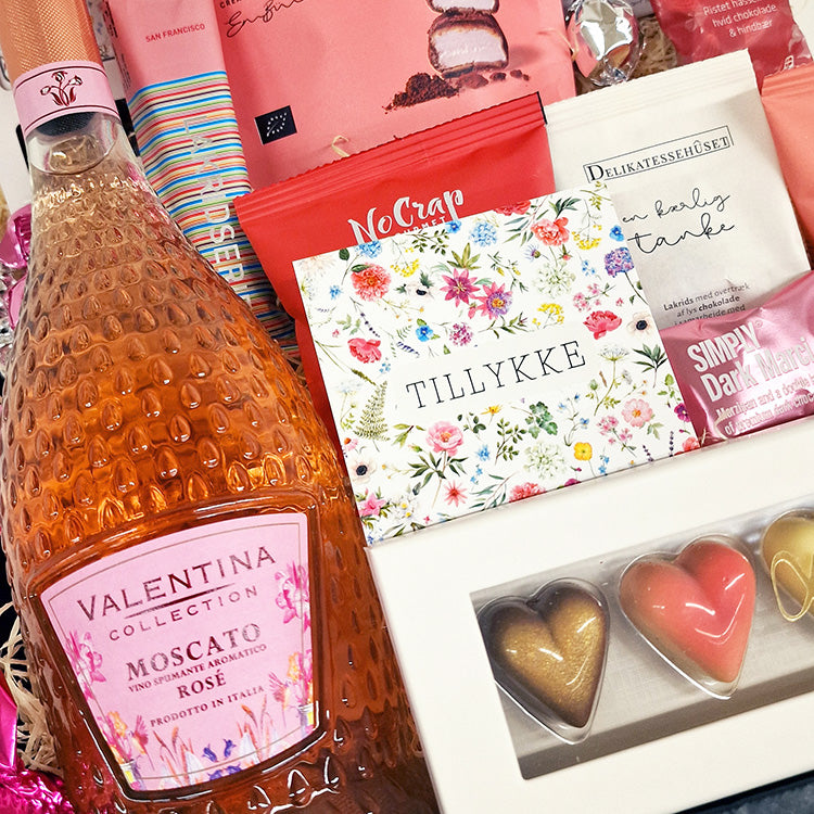 Lyserød gavekurv med Moscato Rosé, chokoladehjerter og kort med teksten tillykke til fejring.