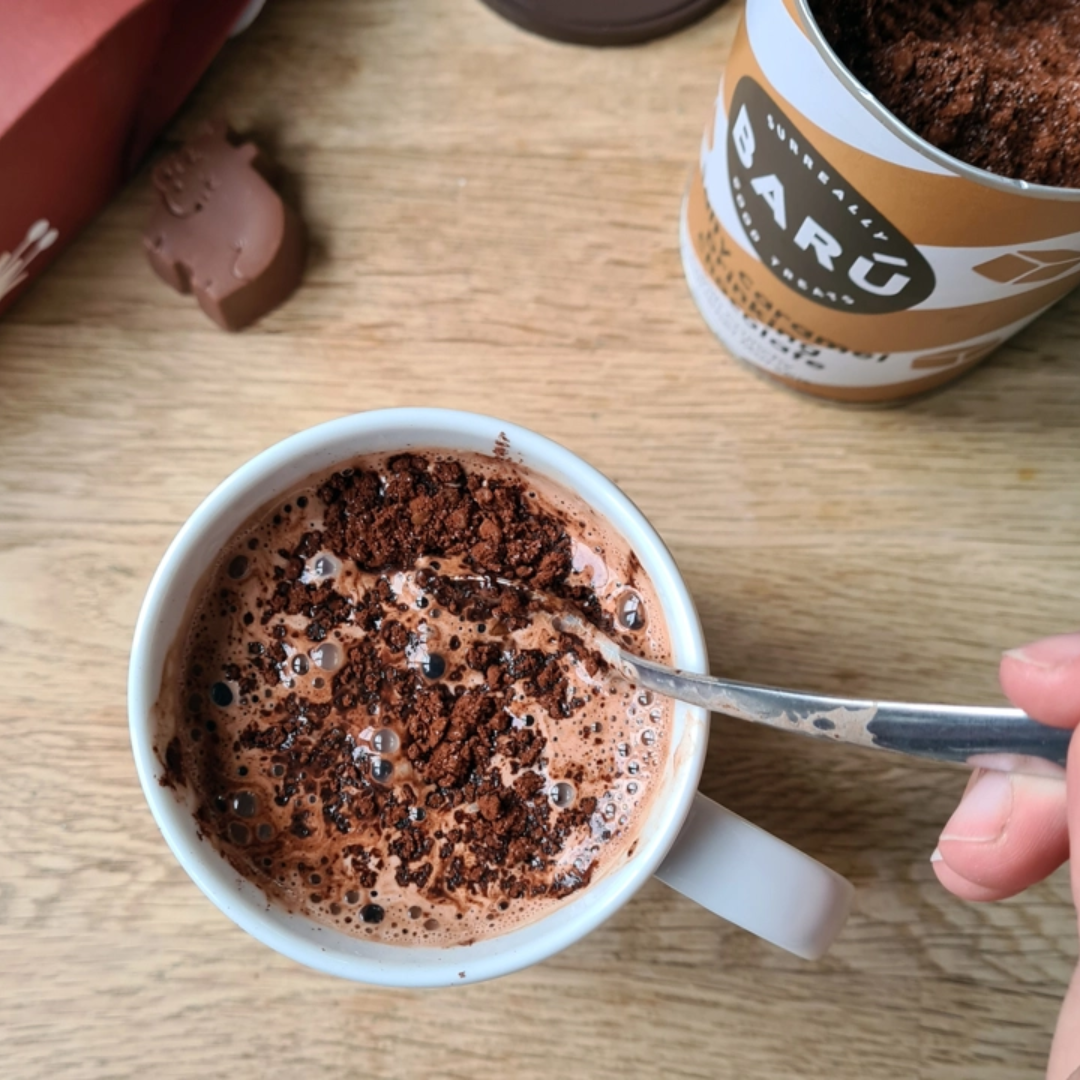 Barú Caramel Hot Chocolate pulver i en kop på et træbord ved siden af dåsen.
